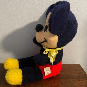 May include: Vintage Mickey Mouse plush toy. The plush features a black body, ears, and head, with a tan face and yellow feet. Mickey wears red shorts and a yellow ribbon. The toy is sitting on a wooden surface.