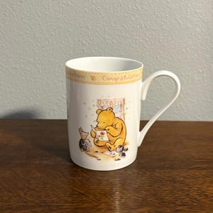 May include: White ceramic mug featuring Winnie the Pooh and Piglet. The mug has a yellow rim with the word "Congratulations" printed on it. The illustration shows Pooh Bear and Piglet. The mug is on a wooden surface.
