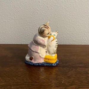 Puede incluir: Figura de cerámica que representa un gato abrazando a otro gato. La figura presenta una paleta de colores rosa, blanco, marrón, amarillo y azul. Los gatos están colocados sobre una base azul con detalles amarillos y naranjas.