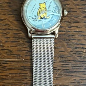May include: Silver-toned wristwatch featuring a light blue face with a Winnie the Pooh illustration. The watch has a mesh band and a classic buckle closure. A charming accessory for fans of the beloved character.