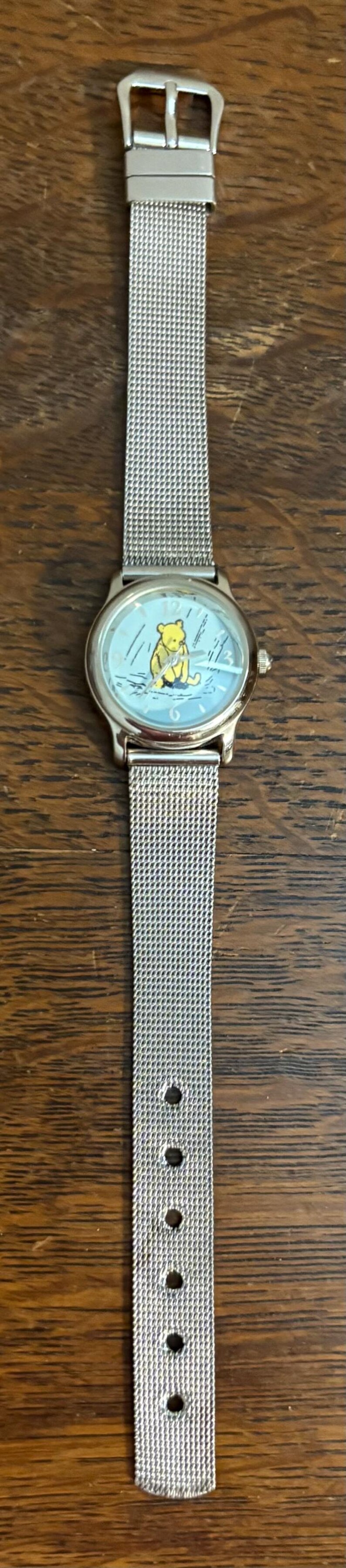 May include: Silver-toned wristwatch featuring a light blue face with a Winnie the Pooh illustration. The watch has a mesh band and a classic buckle closure. A charming accessory for fans of the beloved character.