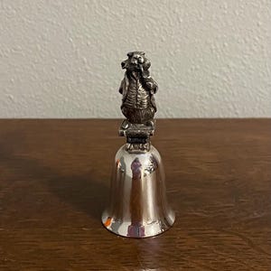 May include: Silver bell with a detailed figurine of a man holding a bunch of flowers on top. The bell is polished and shiny.