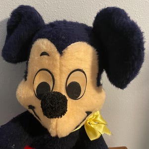 May include: Vintage plush Mickey Mouse toy. The toy features a black and cream-colored face with large black ears. A yellow bow is tied around the neck. The toy has a red heart-shaped detail on the chest.