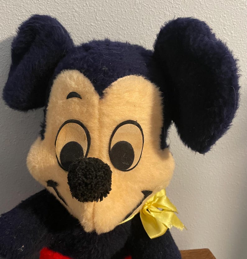 May include: Vintage plush Mickey Mouse toy. The toy features a black and cream-colored face with large black ears. A yellow bow is tied around the neck. The toy has a red heart-shaped detail on the chest.