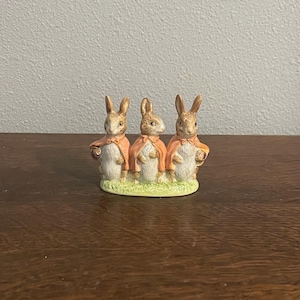 May include: A ceramic figurine featuring three rabbits, each wearing a peach-colored cape. The rabbits stand on a green base, with detailed facial features and holding baskets. The figurine is a decorative item.