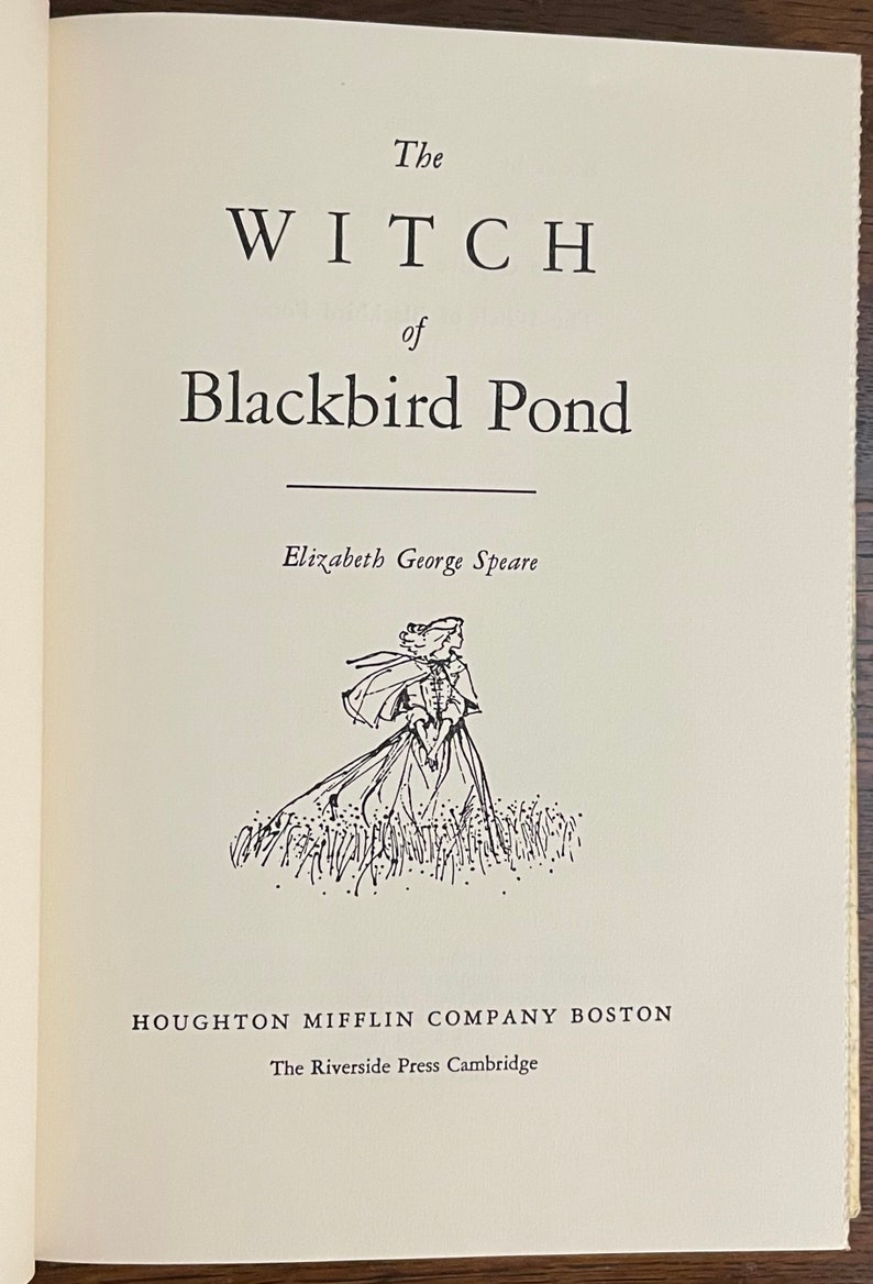 May include: The cover of a vintage book titled "The Witch of Blackbird Pond" by Elizabeth George Speare. The book cover features a black and white illustration of a woman in a long dress and cloak walking through a field of tall grass.