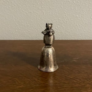 May include: Silver bell with a cat figurine holding a plate and a pitcher. The cat is wearing a dress and a hat.