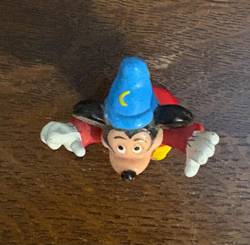May include: A Mickey Mouse figurine in a sorcerer's outfit, featuring a blue hat with a yellow crescent moon, a red robe, and white gloves. The figure is lying on a wooden surface.