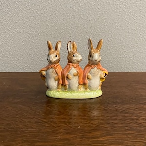 May include: Three brown and white ceramic rabbits wearing orange capes stand on a green base. The rabbits are facing forward and have black eyes and noses.