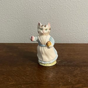 Puede incluir: Una figura de cerámica de un gato con un vestido blanco y azul claro, sosteniendo un pequeño objeto amarillo. El gato tiene ojos verdes y una cabeza estampada. La figura está sobre una superficie de madera.