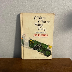 May include: A vintage copy of "Chitty Chitty Bang Bang: The Magical Car" by Ian Fleming, illustrated by John Burningham. The book cover features a green car illustration and the title in a whimsical font. The book is on a wooden surface.