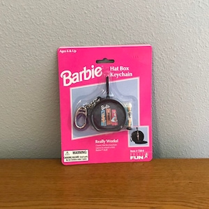 Può includere: Una confezione rosa e bianca con un portachiavi Barbie Hat Box. Il portachiavi è una scatola di cappello nera con una catena argentata e una piccola bambola Barbie all'interno. La confezione riporta la scritta "Really Works!" e "Item # 728-0".