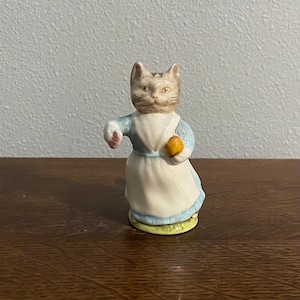 Puede incluir: Una figura de cerámica de un gato vestido con un vestido azul y blanco, sosteniendo un pequeño objeto naranja. El gato tiene una cara detallada con pelaje marrón y canela. La figura está sobre una base verde y amarilla.