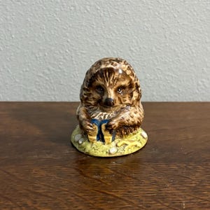 May include: A small ceramic hedgehog figurine with brown and tan textured quills, a dark nose, and dark eyes. The hedgehog is sitting on a light green base with small white accents and is holding a small blue object between its front paws.