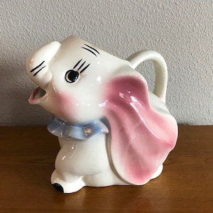 May include: A white ceramic pitcher shaped like an elephant with pink ears and a blue bow. The elephant has black eyes and a black mouth.