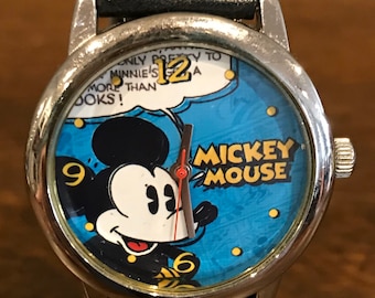 Könnte beinhalten: Eine silberne Mickey Mouse Uhr mit einem schwarzen Lederarmband. Das Zifferblatt der Uhr zeigt ein Cartoon-Bild von Mickey Mouse mit den Worten "Mickey Mouse" in gelben Buchstaben. Das Zifferblatt der Uhr hat auch eine Sprechblase mit dem Text "Only pretty to Minnie's 12 a more than looks!"