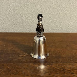 May include: A silver-colored bell with a detailed figure on top. The bell has a flared base and a smooth, reflective surface. The figure appears to be a person wearing a hat and cloak. The bell is displayed on a wooden surface.