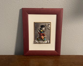 Può includere: Scultura a rilievo in metallo incorniciata di un personaggio dei cartoni animati con giacca rossa e cappello nero. L'opera è ambientata su un passe-partout color crema e incorniciata in una cornice di legno rosso scuro. La scultura è su una superficie di legno.