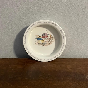 Puede incluir: Plato de cerámica blanca con una colorida ilustración de un conejo y un hombre en una escena de jardín. El plato tiene texto alrededor del borde. Una pieza encantadora para servir o exhibir.