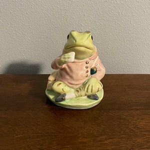 Puede incluir: Una figurita de cerámica de una rana con una chaqueta rosa y zapatos negros, sentada sobre una base verde. La rana sostiene un pequeño objeto blanco en su mano.