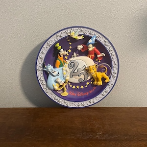 May include: A commemorative plate for the 25th anniversary of Walt Disney World. The plate features embossed Disney characters such as Mickey Mouse, Goofy, and Simba, set against a purple backdrop with gold stars and the text "Walt Disney World".