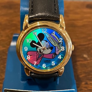 May include: A gold-tone Mickey Mouse watch with a black leather band. The watch face features a colorful iridescent design with Mickey Mouse wearing a hat and a suit. The watch face also includes the text "Lorus Quartz" and "Disney".
