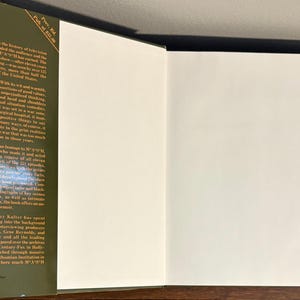 Op de afbeelding: Open boek met een groene kaft en witte pagina's. De linkerpagina heeft zwarte tekst, inclusief de titel en de naam van de auteur. De rechterpagina is leeg. Het boek ligt op een houten oppervlak.