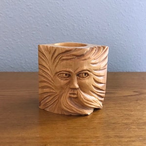 Könnte beinhalten: Ein Kerzenhalter aus Holz, der in Form eines Gesichts mit Bart und windgepeitschtem Haar geschnitzt ist. Der Halter ist hellbraun und hat eine quadratische Form.