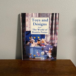 May include: A book titled "Toys and Designs from The World of Beatrix Potter" with a blue and white toy chest on the cover. The chest is filled with stuffed animals and toys. The book is published by F. Warne & Co.