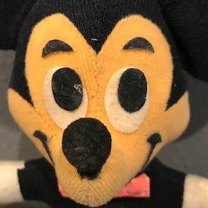 May include: Close-up of a vintage Mickey Mouse plush toy. The toy features a black head with large, white-rimmed eyes, a black nose, and a smiling mouth. The face is a light tan colour. The toy is wearing a black shirt with a red bow tie.