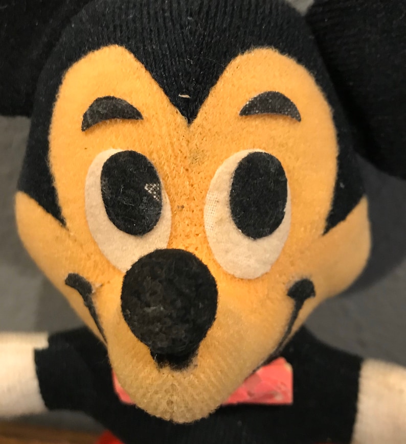 May include: Close-up of a vintage Mickey Mouse plush toy. The toy features a black head with large, white-rimmed eyes, a black nose, and a smiling mouth. The face is a light tan colour. The toy is wearing a black shirt with a red bow tie.