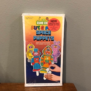 May include: A Sesame Street themed craft kit featuring four stick puppets with reusable vinyl pieces. The puppets are dressed as astronauts and robots. The box features the text "Put & Play Space Puppets".