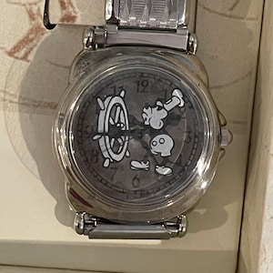May include: A silver wristwatch with a black face and a Mickey Mouse character holding a steering wheel. The watch has a silver band with a textured design.
