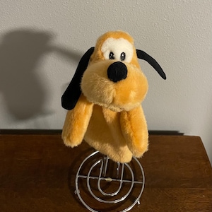 Puede incluir: Un títere de perro de peluche marrón y negro con nariz negra y ojos grandes negros. El títere está de pie sobre un soporte de metal.