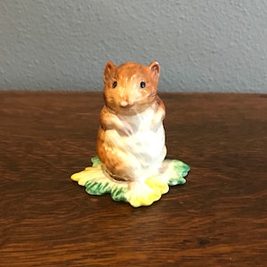 May include: A small ceramic figurine of a brown mouse sitting on a green and yellow leaf. The mouse has black eyes and a white belly.