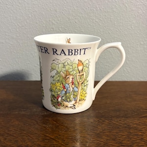 Peut inclure: Tasse en céramique blanche avec une illustration de Pierre Lapin. La tasse a une anse incurvée et un motif coloré de Pierre Lapin tenant des carottes, avec l'inscription "PETER RABBIT" sur le côté. La tasse est posée sur une surface en bois.