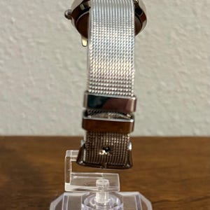 May include: A silver wristwatch with a mesh band is displayed on a clear stand. The watch has a round face and a metallic band. The watch is on a clear plastic stand with the text "MADE IN CHINA" visible.