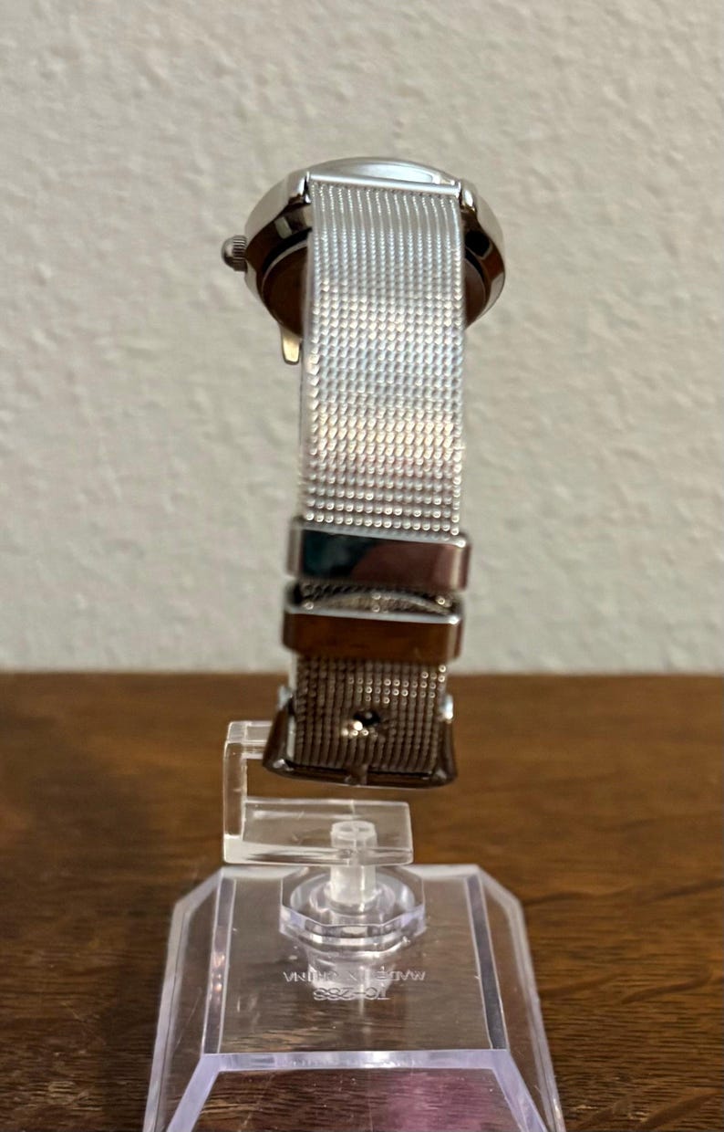 May include: A silver wristwatch with a mesh band is displayed on a clear stand. The watch has a round face and a metallic band. The watch is on a clear plastic stand with the text "MADE IN CHINA" visible.