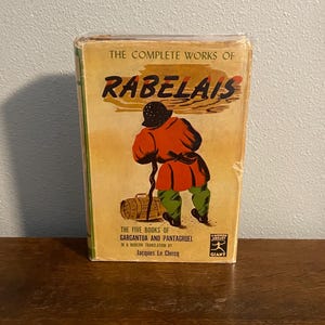 Peut inclure: Un livre ancien intitulé "The Complete Works of Rabelais". La couverture présente une illustration d'une personne en manteau rouge et pantalon vert, portant un bâton et un tonneau. Le livre fait partie de la série Modern Library Giant.
