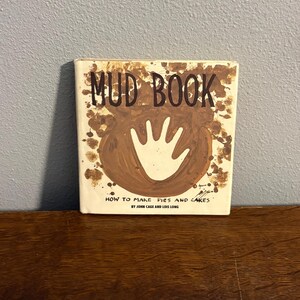 May include: A square hardcover book titled Mud Book: How to Make Pies and Cakes by John Cage and Lois Long. The cover is off-white with a large brown circular mud splatter design in the center, featuring a white handprint silhouette.