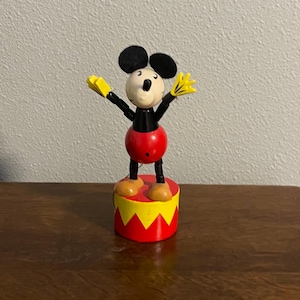 May include: A vintage wooden Mickey Mouse puppet with black ears, red shorts, and yellow gloves. The puppet is attached to a red and yellow base.