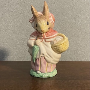 Puede incluir: Una figurita de cerámica de un conejo que lleva un vestido rosa y blanco y una capa roja con capucha. El conejo sostiene un paraguas verde y una cesta de mimbre con huevos blancos.