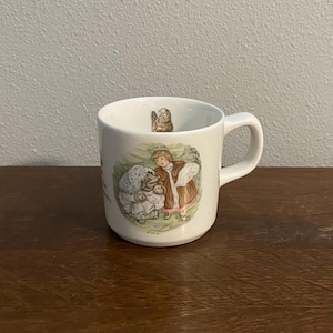 May include: White ceramic mug with a handle, featuring a colorful illustration of two figures in a pastoral scene. The mug has a small illustration inside the rim. The mug is sitting on a wooden surface.