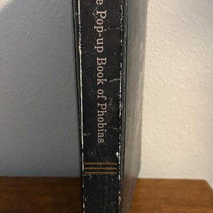 May include: A hardcover book titled "The Pop-up Book of Phobias" with a black cover and white text. The book is standing upright on a wooden surface, with the spine facing the viewer.