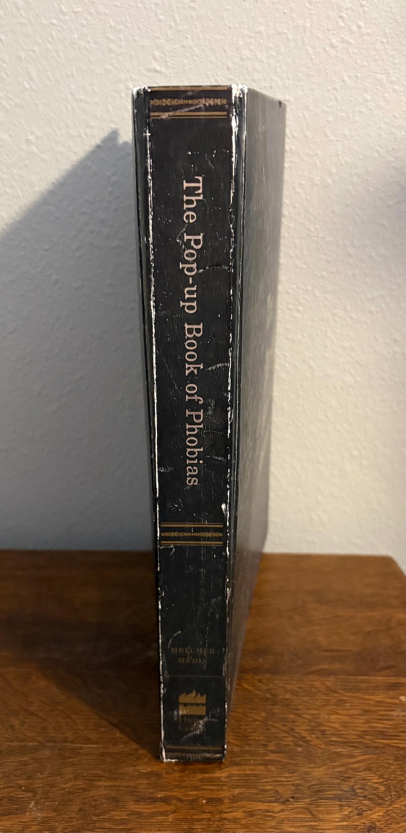 May include: A hardcover book titled "The Pop-up Book of Phobias" with a black cover and white text. The book is standing upright on a wooden surface, with the spine facing the viewer.