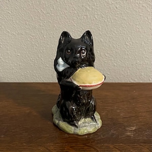 May include: A black ceramic figurine of a dog holding a pie. The dog has a white bow tie and is sitting on a green and brown base. The pie has a yellow top with a red rim.
