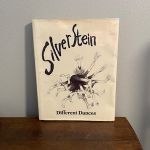 May include: A white book with the title "SilverStein" in black cursive lettering. The cover features a black and white illustration of a person with multiple arms and legs dancing. The text "Different Dances" is printed in black at the bottom of the cover.