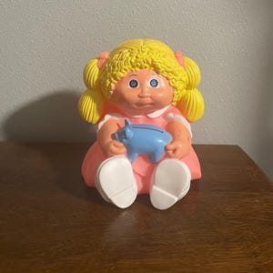 Pode incluir: Um cofrinho vintage Cabbage Patch Kids em forma de boneca. A boneca tem cabelo loiro encaracolado, olhos azuis e usa um vestido rosa. Ela está segurando um cofrinho azul. O cofrinho está em uma superfície de madeira.