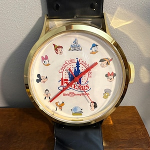 May include: A large wall clock designed like a wristwatch with a gold-tone metal frame and a black strap. The white face features illustrations of Disney characters, a castle, and text reading Coca-Cola Celebrates 15 Years Walt Disney World.