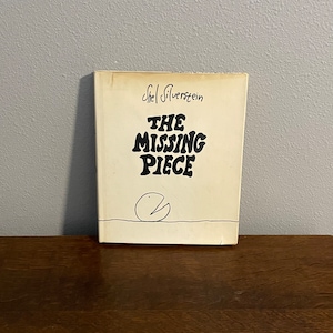 May include: A book titled "The Missing Piece" by Shel Silverstein. The cover is off-white with black text and a simple line drawing of a Pac-Man-like shape. The book is on a wooden surface.